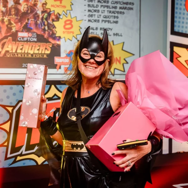 A woman in a Batgirl costume smiles, holding a pink-wrapped bouquet and a gift box, with comic book-themed artwork and an Avengers poster in the background.