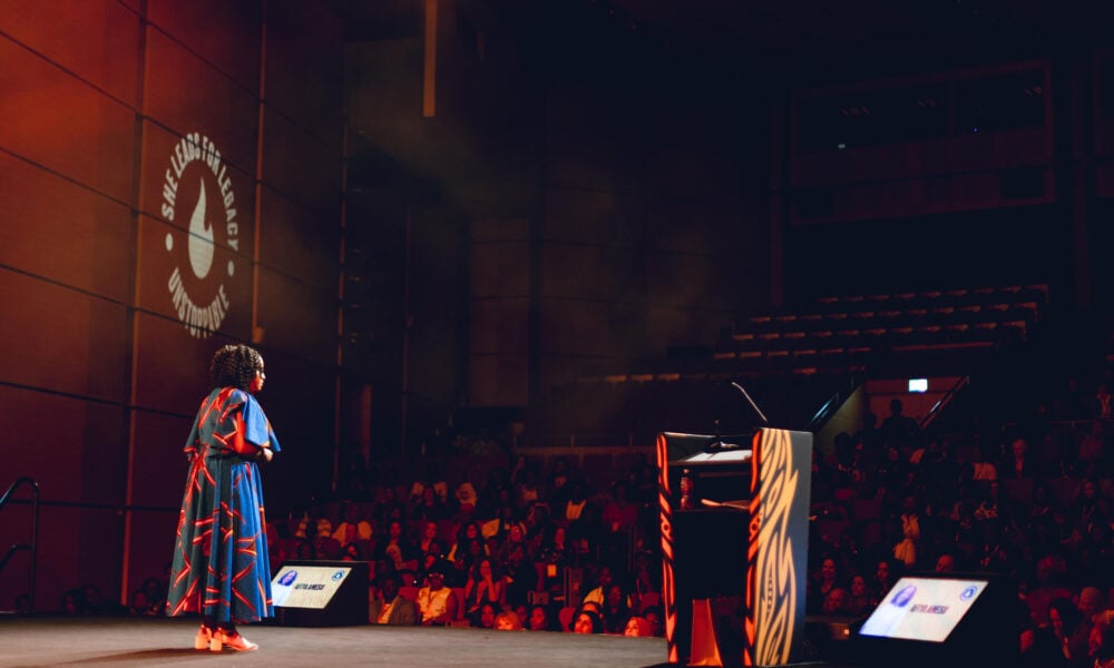 A person in a colourful dress stands on stage facing an audience in a dimly lit auditorium, with a lectern and large screens nearby, and event branding projected on the wall.