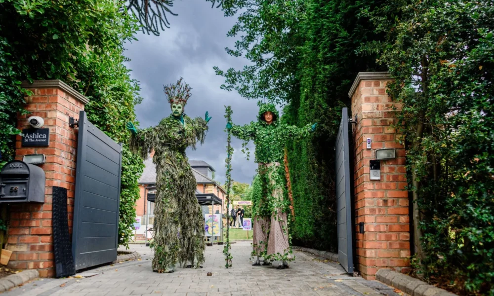 Two people in tall costumes covered in green foliage stand on stilts at the entrance of a brick-walled drive, beneath a cloudy sky, with trees and a house visible in the background.