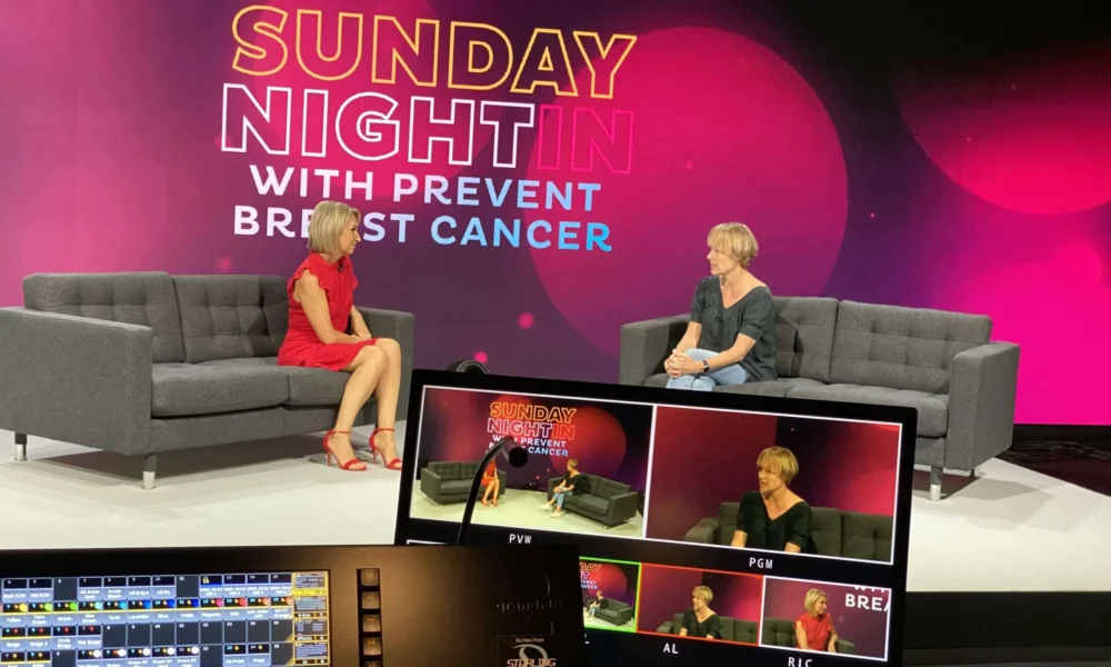 Two women sit on stage, chatting on grey sofas for a Sunday Night In with Prevent Breast Cancer event. A monitor in the foreground displays multiple live camera feeds of the discussion.