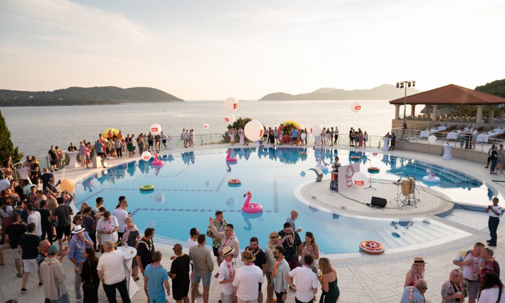 Large crowd gathers around a pool with inflatable toys at a waterfront venue during sunset, with mountains and the sea in the background. Some people chat, whilst others enjoy the festive atmosphere.