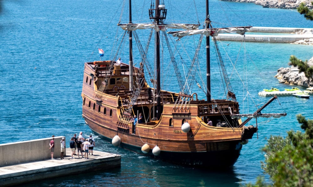 A large wooden sailing ship docks near a concrete quay with people standing nearby, surrounded by clear blue water and distant green hills.