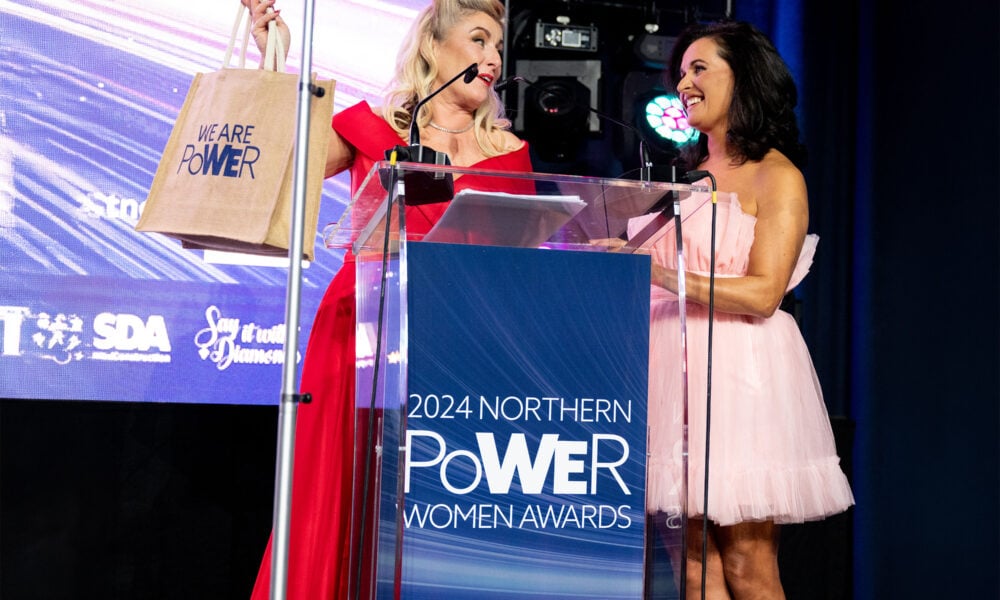 Two women in dresses stand at a lectern labelled 2024 Northern Power Women Awards. One woman holds a We Are Power bag, smiling at the other. Bright stage lighting and event sponsor logos appear in the background.